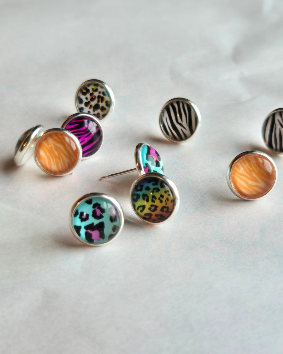 A group of round stud earrings with silver-toned bezel settings and colorful animal print and zebra-patterned cabochons, arranged on a light neutral surface.