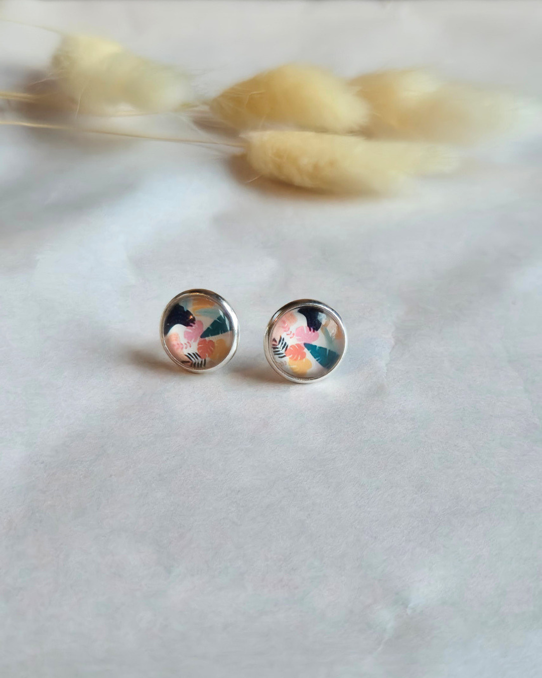 A pair of silver stud earrings with colorful abstract art designs, resting on a white textured surface with blurred dried grass in the background.