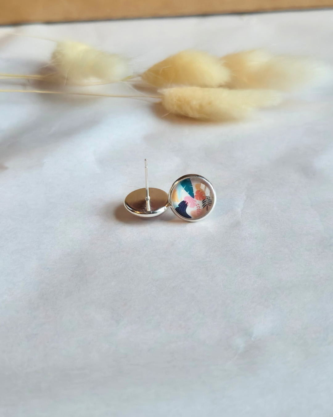 A pair of small round stud earrings with colorful abstract artwork on a clear dome, shown from both front and back on a white textured surface with soft dried grass in the background.
