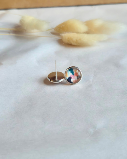 A pair of small round stud earrings with colorful abstract artwork on a clear dome, shown from both front and back on a white textured surface with soft dried grass in the background.