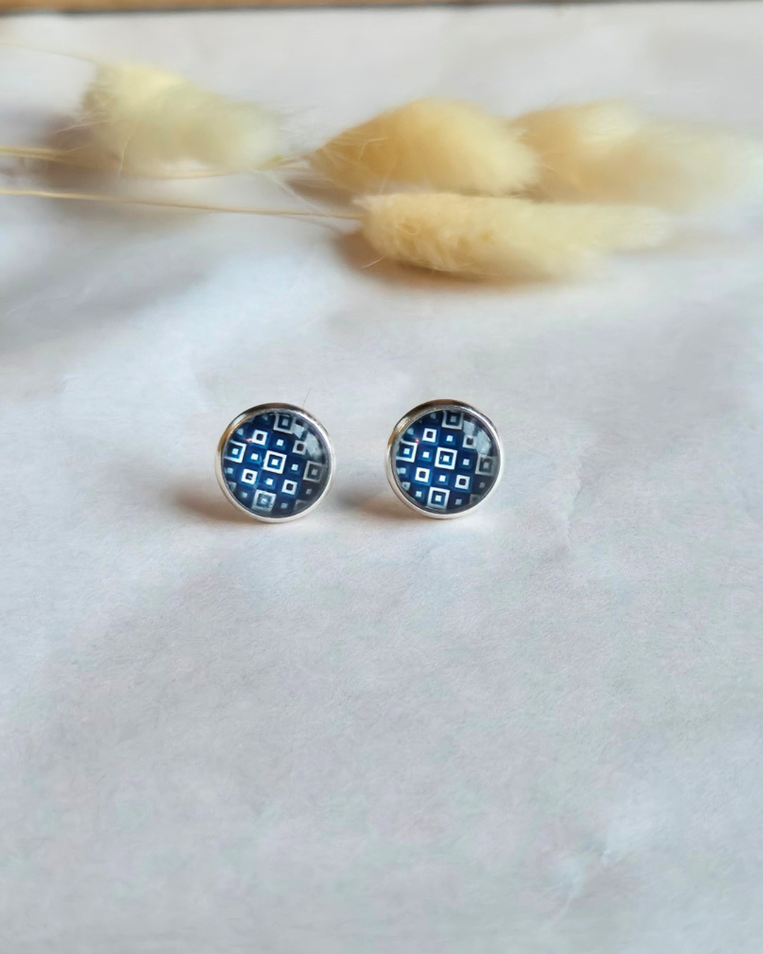 A pair of round stud earrings with a blue and white geometric pattern, set in a silver-toned metal, displayed on a white textured surface with dried bunny tail grass in the background.