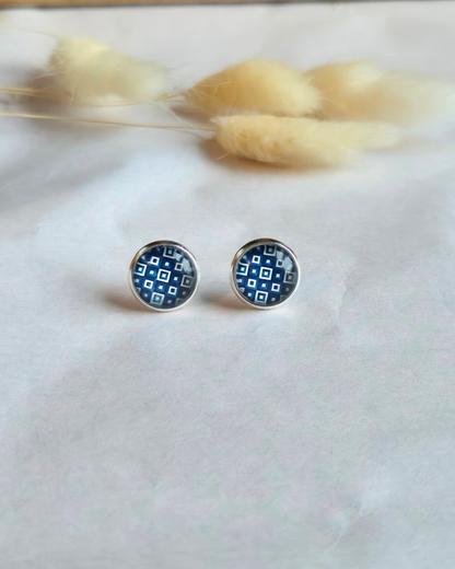 A pair of round stud earrings with a blue and white geometric pattern, set in a silver-toned metal, displayed on a white textured surface with dried bunny tail grass in the background.