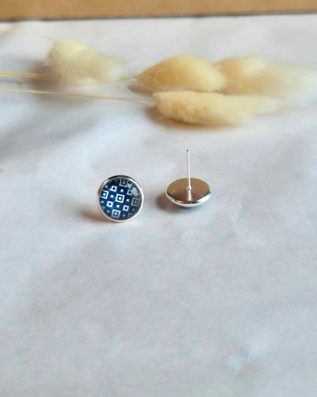 A pair of small round stud earrings displayed on a light surface with dried grass stems in the background. One earring is shown from the front, featuring a blue and white geometric pattern inside a silver-toned bezel, and the other is shown from the back with a silver post and flat back.