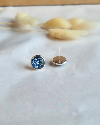 A pair of small round stud earrings displayed on a light surface with dried grass stems in the background. One earring is shown from the front, featuring a blue and white geometric pattern inside a silver-toned bezel, and the other is shown from the back with a silver post and flat back.