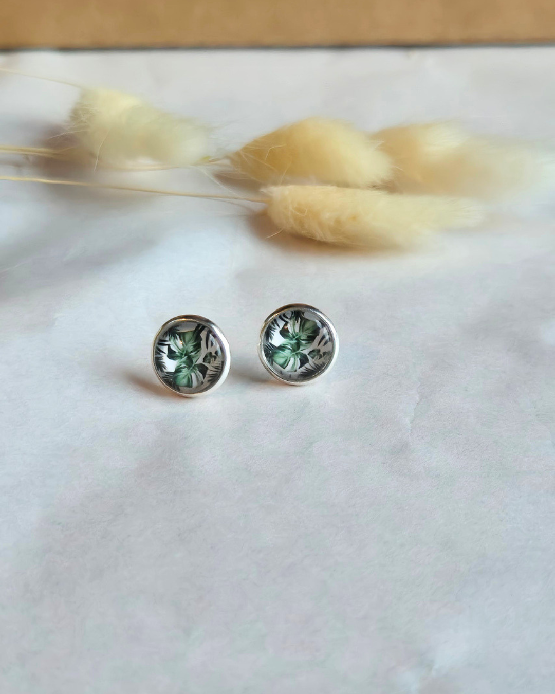 A pair of round stud earrings with green floral designs set in silver, displayed on a white textured surface with dried bunny tail grass in the background.