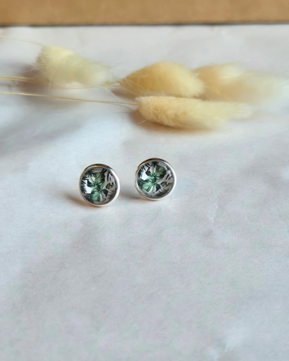 A pair of round stud earrings with green floral designs set in silver, displayed on a white textured surface with dried bunny tail grass in the background.