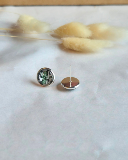 A pair of round stud earrings shown on a light surface with dried grass stems in the background. One earring is facing forward, showing a silver-toned bezel with a green and black marbled or textured inlay.