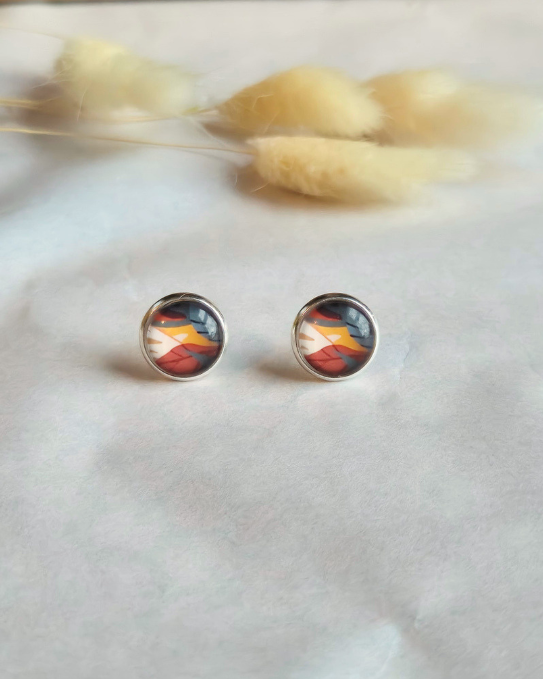 A pair of round stud earrings with a silver-toned bezel and a colorful abstract design featuring red, orange, yellow, and dark blue/black tones, displayed on a light textured surface with soft dried grass in the background.