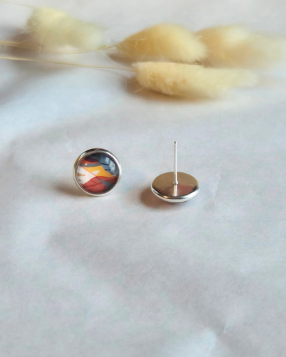 A pair of silver stud earrings with colorful abstract art domes, shown on a soft white textured fabric with dried bunny tail grass in the background.