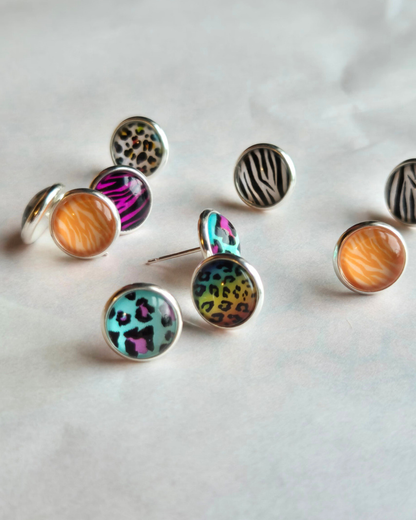 A group of round stud earrings with silver-toned bezel settings and colorful animal print and zebra-patterned cabochons, arranged on a light neutral surface.