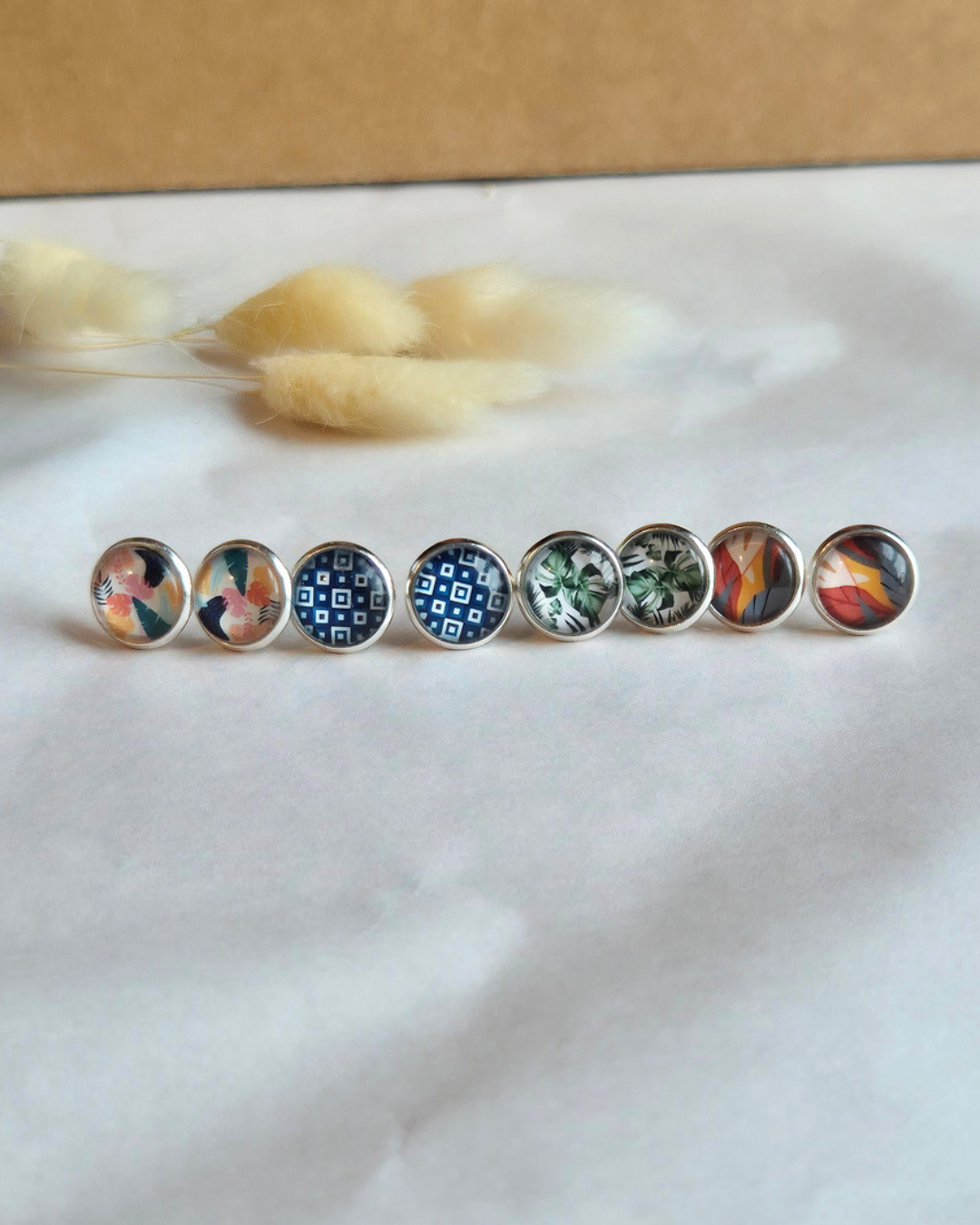 Eight round stud earrings with colorful abstract and patterned designs in silver-toned settings, arranged in a row on a white textured surface with dried grass stems in the background.